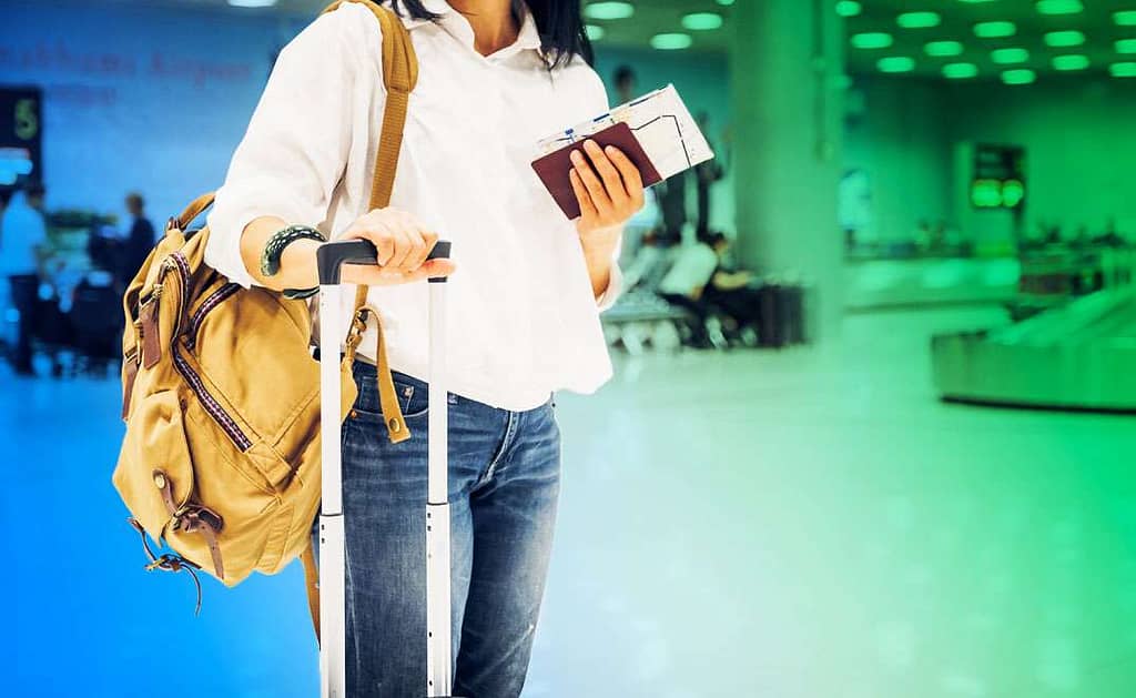 A person at the airport holding their passport and ticket, and carrying their backpack and suitcase.