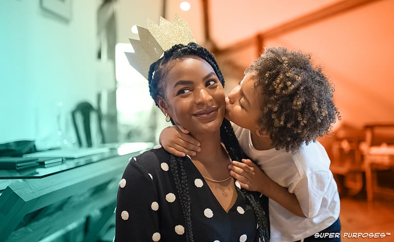A smiling mom is wearing a paper crown and getting a kiss on the cheek from their child.